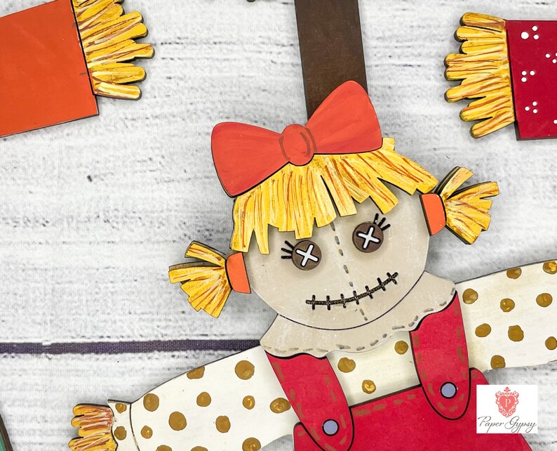 May include: A wooden cutout of a scarecrow with a red and white polka dot dress, a red bow, and a straw hat. The scarecrow has a stitched smile and button eyes.