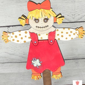 May include: A wooden cutout of a rag doll scarecrow with a red dress, yellow hair, and a red bow. The scarecrow has a stitched smile and X-shaped eyes. The scarecrow is holding out its arms with white and gold polka dot sleeves.