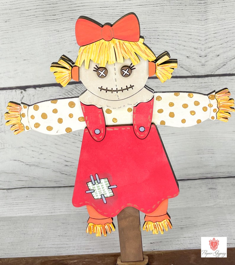 May include: A wooden cutout of a rag doll scarecrow with a red dress, yellow hair, and a red bow. The scarecrow has a stitched smile and X-shaped eyes. The scarecrow is holding out its arms with white and gold polka dot sleeves.