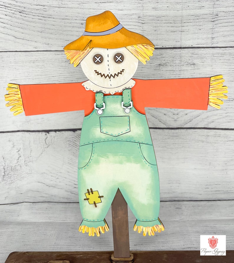 May include: A friendly scarecrow with a straw hat, orange shirt, and blue overalls. The scarecrow has a stitched-on smile and X's for eyes. The scarecrow is standing on a wooden stake.