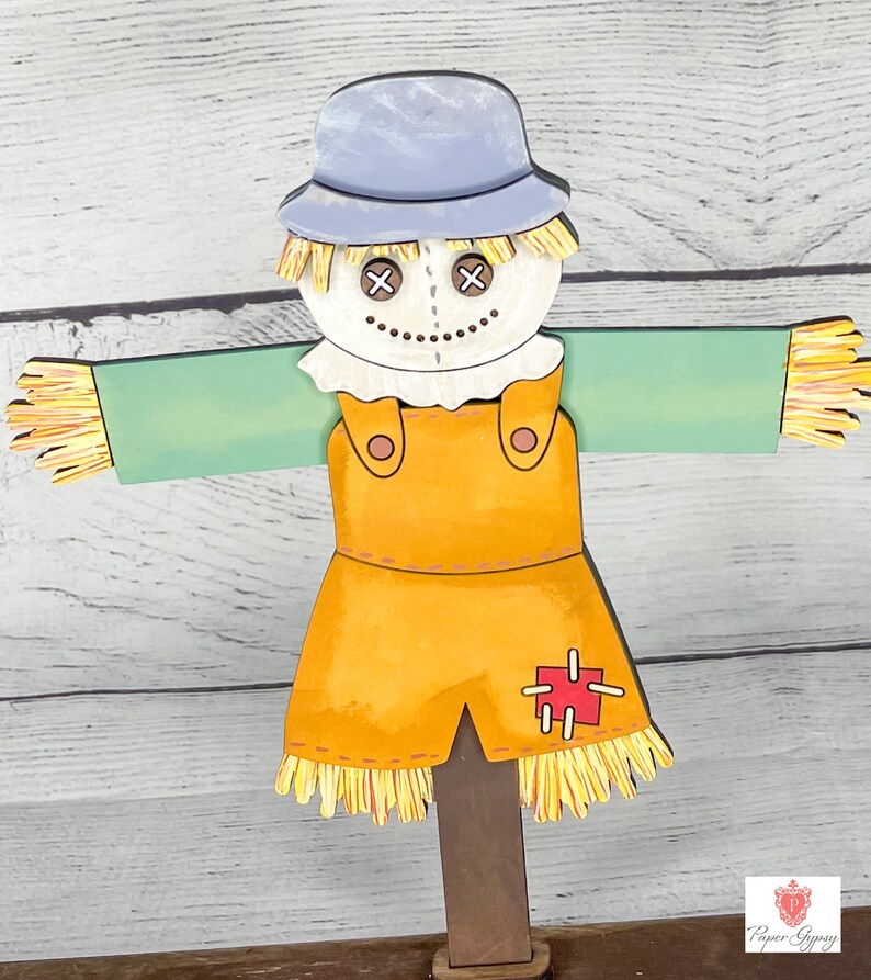 May include: A wooden scarecrow with a blue hat, white face, and orange overalls. The scarecrow has a red patch on the overalls and straw accents on the arms and legs.