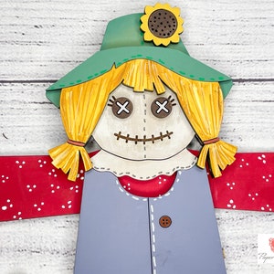 May include: A wooden cutout of a scarecrow with a green hat, yellow hair, and a red and white checkered shirt. The scarecrow has a stitched-on face with X-shaped eyes and a stitched mouth.