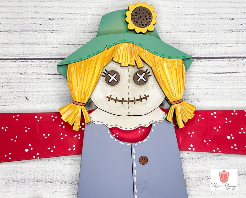 May include: A wooden cutout of a scarecrow with a green hat, yellow hair, and a red and white checkered shirt. The scarecrow has a stitched-on face with X-shaped eyes and a stitched mouth.