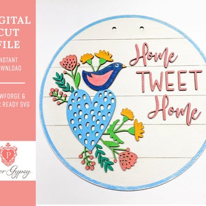 Puede incluir: Un letrero redondo de madera con un borde azul y un fondo blanco. El letrero presenta un pájaro azul con el vientre rosa sentado en un corazón azul con lunares blancos. El corazón está rodeado de hojas verdes y flores rosas y amarillas. El texto "Home Tweet Home" está escrito en letras rosas en el letrero.