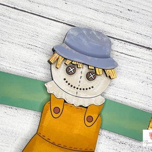 May include: A wooden scarecrow with a blue hat, brown overalls, and a straw-like beard. The scarecrow has a painted face with brown eyes and a stitched smile. The scarecrow is holding out its arms, which are painted green.
