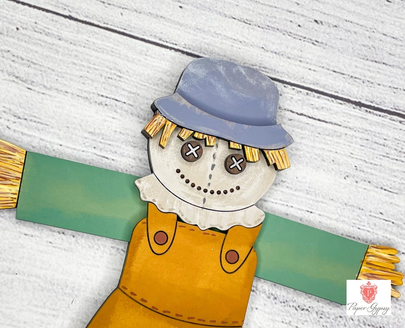 May include: A wooden scarecrow with a blue hat, brown overalls, and a straw-like beard. The scarecrow has a painted face with brown eyes and a stitched smile. The scarecrow is holding out its arms, which are painted green.
