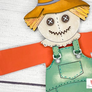 May include: A wooden scarecrow cutout with a straw hat, orange shirt, and green overalls. The scarecrow has a stitched-on face with brown eyes and a stitched-on smile.