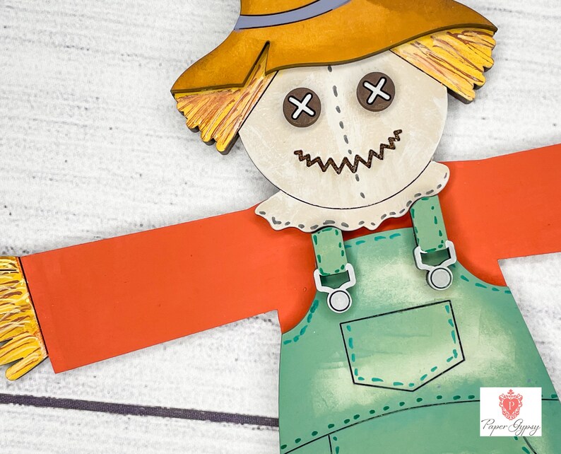 May include: A wooden scarecrow cutout with a straw hat, orange shirt, and green overalls. The scarecrow has a stitched-on face with brown eyes and a stitched-on smile.
