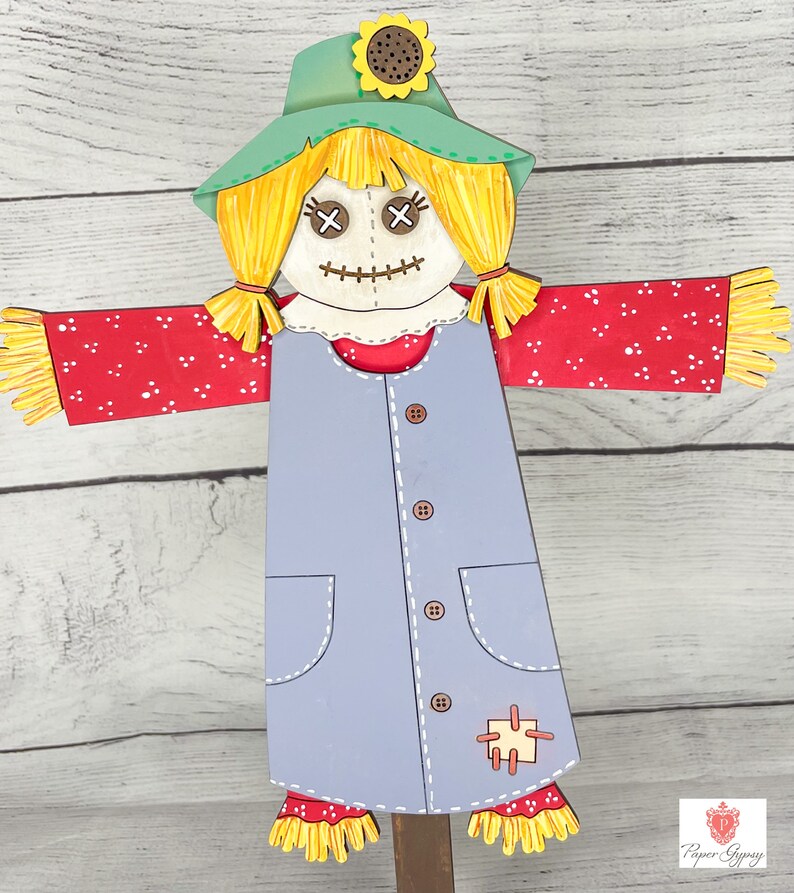 May include: A wooden scarecrow with a green hat, yellow hair, red and white polka dot sleeves, and a blue dress with buttons and pockets. The scarecrow has a stitched smile and X-shaped eyes.