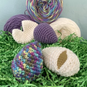 May include: A collection of crochet items including a multi-colored yarn ball, a solid purple yarn ball, and a solid white yarn ball. There are also two small, egg-shaped crochet pieces, one in multi-colored yarn and one in solid white yarn.