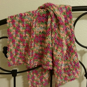 May include: A crocheted shawl in a multi-coloured pattern of pink, green, and purple. The shawl is draped over a black metal bed frame.