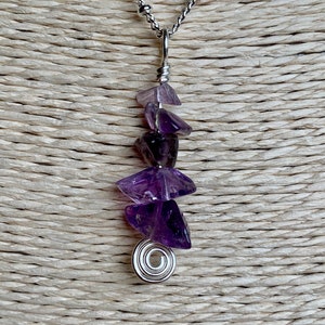 May include: A silver chain necklace with a pendant made of multiple purple amethyst stones. The stones are arranged in a cascading design and are held together by silver wire. The pendant is finished with a silver spiral.