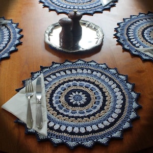 May include: A set of four crocheted placemats in shades of blue, beige and white. The placemats feature a circular mandala design with intricate details. A silver tray with salt and pepper shakers sits on top of one of the placemats.