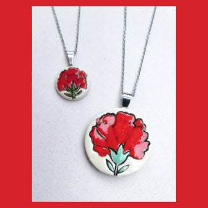 May include: Two silver necklaces with red carnation flower pendants. The larger pendant is a detailed illustration of a red carnation with green leaves and a white background. The smaller pendant is a simpler design with a red carnation on a white background.