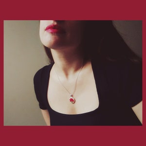 May include: A woman wearing a black scoop neck top with a silver chain necklace and a red pendant. The pendant is a small oval shape with a red flower design.