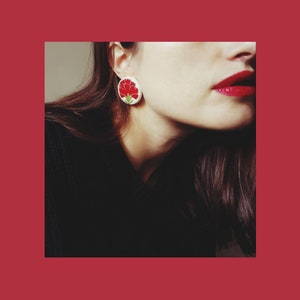 May include: A close-up of a woman's face with a red carnation flower earring. The earring is white with a red carnation flower design.