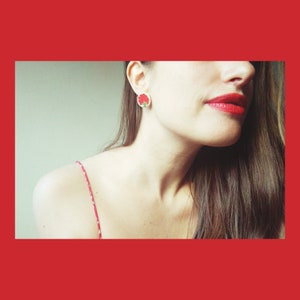 May include: A woman with long brown hair wearing a red and white patterned strap top and a red flower earring.