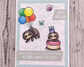 May include: A handmade birthday card featuring a cartoon sloth with colorful balloons and another on a cake. The card has the text "Happy Birthday" and "Let's Pretend This Card Is on Time. MY SPIRIT ANIMAL IS A SLOTH."