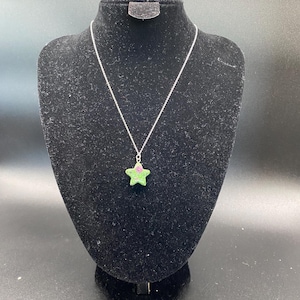 May include: A star-shaped pendant necklace. The pendant is green with a pink accent and hangs from a silver chain. The necklace is displayed on a black velvet bust.