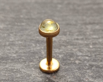 Könnte beinhalten: Ein goldener Körperschmuck mit einem durchsichtigen, grünen Edelstein oben. Das Piercing ist im Labret-Stil, mit einer flachen Basis und einem Stift, der durch die Lippe oder ein anderes Piercing geht.