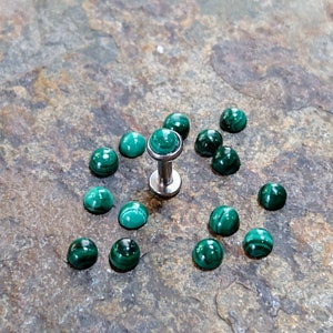 May include: A collection of 14 green malachite gemstone cabochons, each with a unique pattern. The cabochons are arranged around a silver body jewellery post with a malachite top.