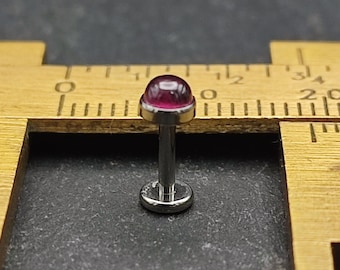 May include: A close-up of a small, pink gemmed body piercing, measured with a ruler. The gem is a dome shape and the post is made of metal.