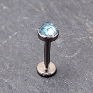 Peut inclure: Un seul piercing pour la lèvre, de couleur argentée, orné d'une gemme bleu clair à facettes. Le bijou a une base plate et une tige qui s'étend jusqu'à la gemme. La gemme est sertie dans une lunette argentée.