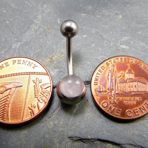 May include: A silver belly button ring with a clear, round gemstone. The ring is shown next to two US pennies for size comparison.