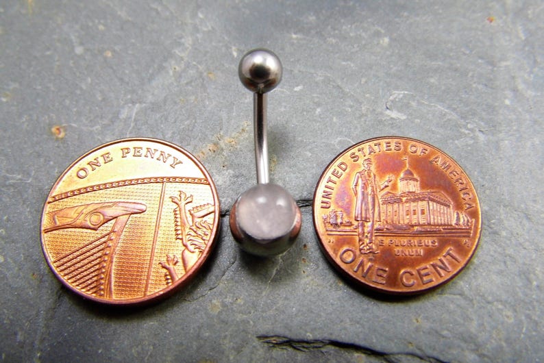 May include: A silver belly button ring with a clear, round gemstone. The ring is shown next to two US pennies for size comparison.
