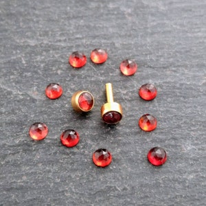 Könnte beinhalten: Ein Set mit 10 roten Edelstein-Steckern mit goldenen Fassungen. Die Stecker sind in einem Kreis um zwei größere Stecker mit goldenen Stiften angeordnet.