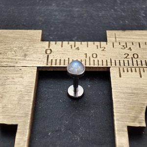 Könnte beinhalten: Ein Körperschmuckstück aus Silber mit einem runden, schillernden Edelstein. Der Schmuck wird an einem Lineal gemessen und zeigt, dass er einen Durchmesser von etwa 10 Millimetern hat.
