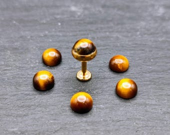 May include: Six gold-toned tiger's eye gemstone beads, one of which is attached to a gold-toned post, a type of body jewelry.