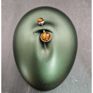 May include: A belly button ring featuring a silver bar and two amber-coloured, round gemstones. The top gem is smaller than the bottom gem. The jewellery is displayed on a green, oval-shaped surface.