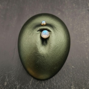 May include: A navel piercing jewellery piece featuring two round, iridescent moonstone cabochons set in gold-toned metal. The larger stone is positioned at the bottom and the smaller stone at the top, both displayed on a textured olive green surface.