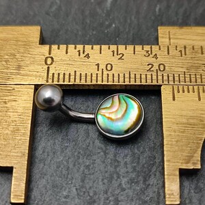 This has an 8mm Paua shell cabochon set in a silver polished surgical steel belly bar. The belly bar ball is 5mm in diameter.

The Paua shell has swirls of colour with green, blue, yellow white and black visible in the pattern.  It is sitting on it's side on a black slate background and next to a brass measuring caliper for both size measurement and contrast.