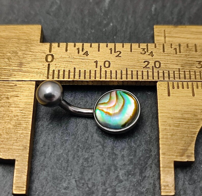 This has an 8mm Paua shell cabochon set in a silver polished surgical steel belly bar. The belly bar ball is 5mm in diameter.

The Paua shell has swirls of colour with green, blue, yellow white and black visible in the pattern.  It is sitting on it's side on a black slate background and next to a brass measuring caliper for both size measurement and contrast.