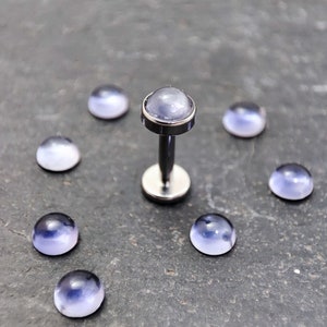 Peut inclure: Un ensemble de huit gemmes en verre transparent avec une nuance irisée violette. Les gemmes sont disposées en cercle autour d'une barre argentée avec une gemme violette sur le dessus. Les gemmes sont destinées à être utilisées pour les bijoux corporels.