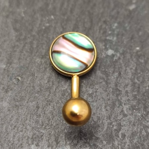 以下が含まれることがあります： 丸いアワビの貝殻のトップが付いた金のボディジュエリー。貝殻は緑、ピンク、黒の虹色の模様をしています。
