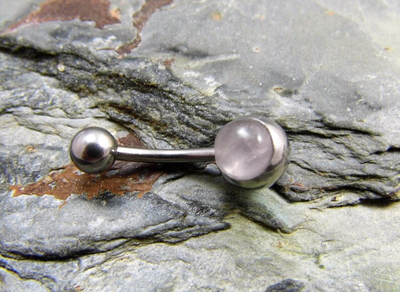 May include: A silver belly button ring with a clear pink gemstone. The ring is on a gray textured surface.