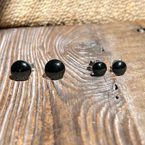 May include: Three pairs of black stud earrings on a wooden surface. The earrings are different sizes.