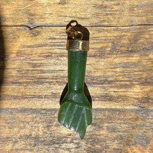 May include: A green jade pendant in the shape of a fist. The pendant has a gold-colored metal cap and bail. The jade is a translucent green color, and the fist is carved with detailed lines. The pendant is displayed on a wooden surface.