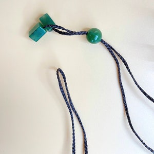 May include: A dark green jade necklace with a round bead and two square beads. The necklace is made of black braided cord.