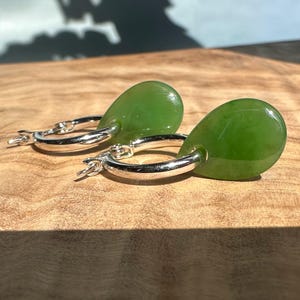 Peut inclure: Boucles d'oreilles créoles en argent avec des pendentifs en forme de goutte de jade vert. Les boucles d'oreilles sont posées sur une surface en bois, mettant en valeur la couleur et la brillance.