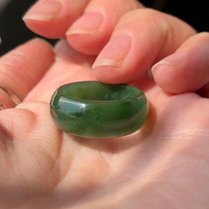 Può includere: Un anello verde giada liscio con una fascia arrotondata. L'anello è di un colore verde opaco uniforme con sottili variazioni di tonalità. L'anello è tenuto in una mano, mostrando la sua superficie lucida e il suo design semplice.