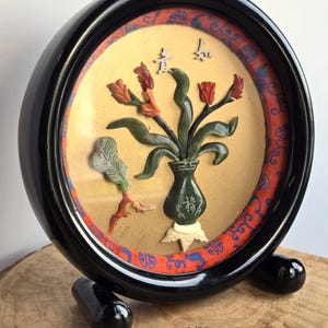 May include: A circular black frame holds a decorative wall art piece featuring a dark green stone vase with a white base and carved stone flowers in shades of red and green. The background is a gold-toned disc surrounded by a red fabric border with blue patterns.