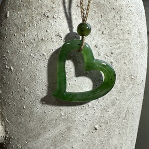 May include: A green jade heart-shaped pendant on a brown cord necklace. The pendant has a smooth, polished surface and a hole in the center.