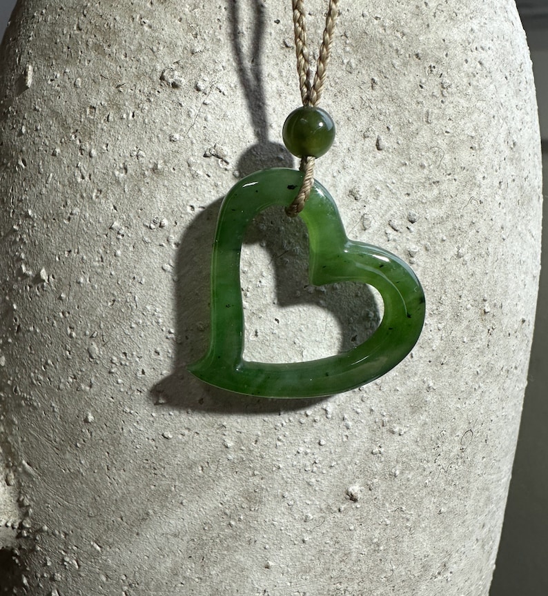 May include: A green jade heart-shaped pendant on a brown cord necklace. The pendant has a smooth, polished surface and a hole in the center.