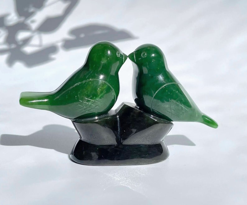Puede incluir: Dos p&aacute;jaros de jade verde tallados en una pose de beso, posados ​​sobre una base de jade negro.