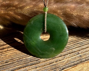 May include: A green jade pendant in the shape of a donut, hanging from a brown cord with a small green bead. The pendant is resting on a wooden surface.