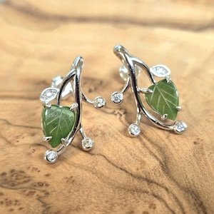 May include: A pair of silver-toned leaf earrings. Each earring features a green leaf-shaped stone, accented with clear, round and oval-shaped crystals. The earrings are set against a wooden background.